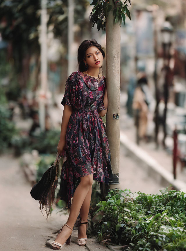 Woman in floral dress and heels with handbag posing by tree in urban setting