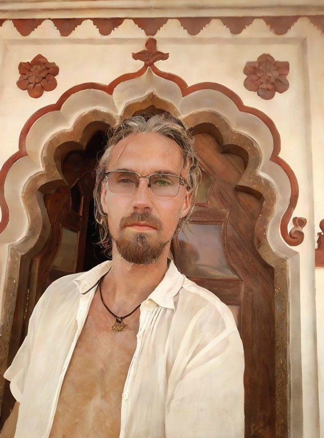 Man with Long Hair and Glasses in Front of Ornamental Wall wearing White Shirt and Necklace