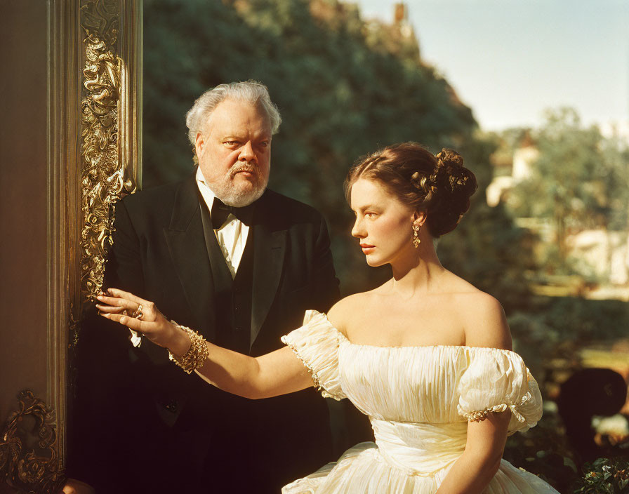Elderly man in tuxedo and young woman in off-shoulder gown by gilded