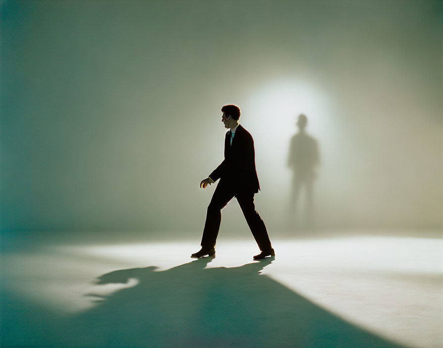 Man walking in lit space with long shadow and looming silhouette in background.
