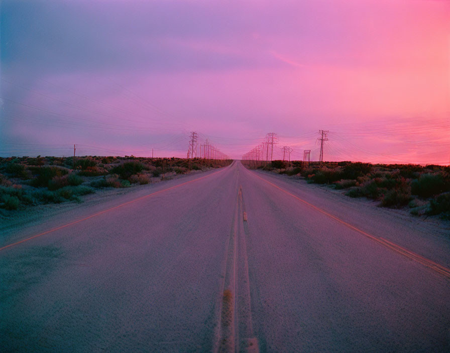 Deserted road under pink and purple dusk sky with power lines and desert vegetation.