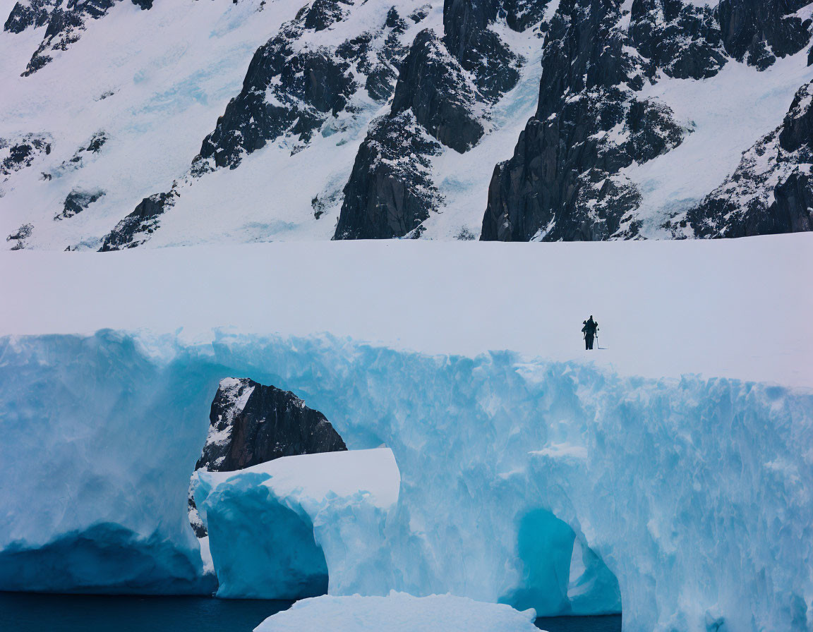 Solitary figure on expansive blue ice arch surrounded by snowy mountains.