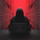 Hooded figure at laptop in red-lit server room with binary code screens