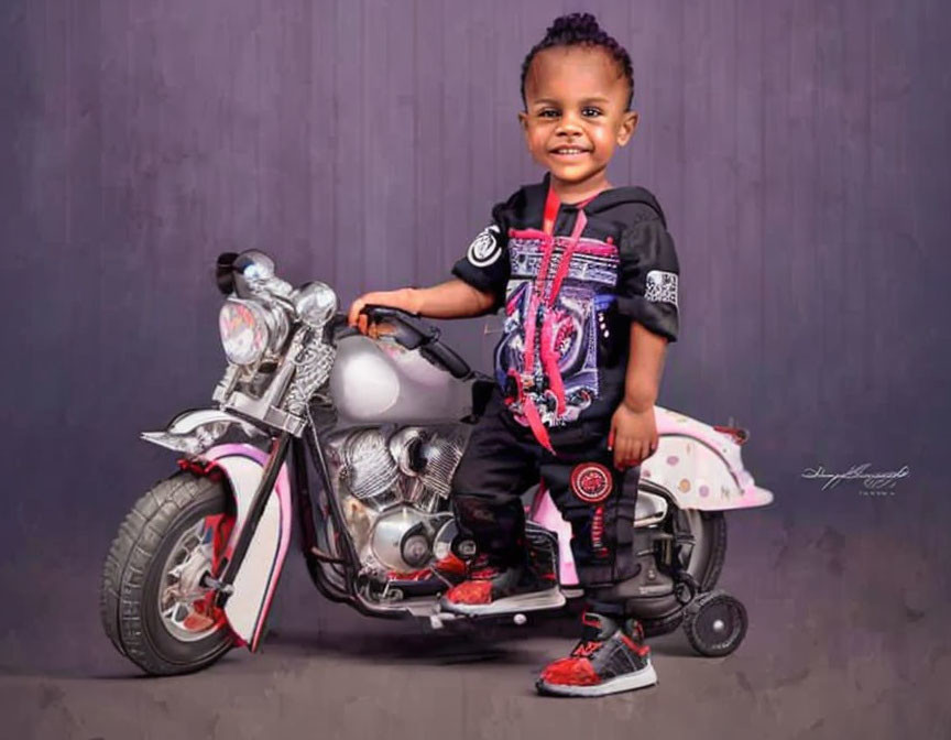 Smiling toddler on toy motorcycle in black and pink outfit on purple background