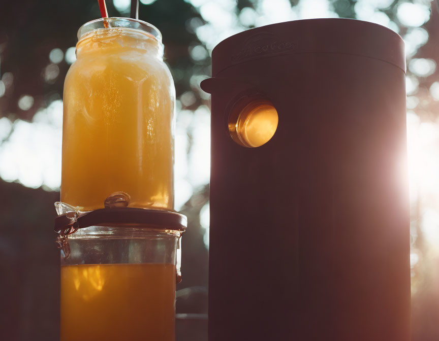 Jar, bottle of juice, and straw near outdoor speaker with sunlight through trees