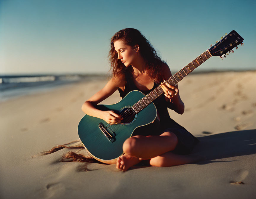 Young woman playing blue guitar on sandy beach with ocean and clear sky