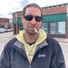 Man in sunglasses smoking cigarette in front of brick building wearing black jacket and yellow hoodie