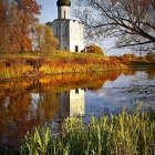 Blue church with golden spire surrounded by autumn trees and calm water.