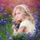 Young girl with blonde curly hair in white dress holding puppy among purple and blue flowers