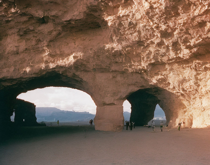 Spacious cave with multiple openings, explorers inside, desert landscape in the background