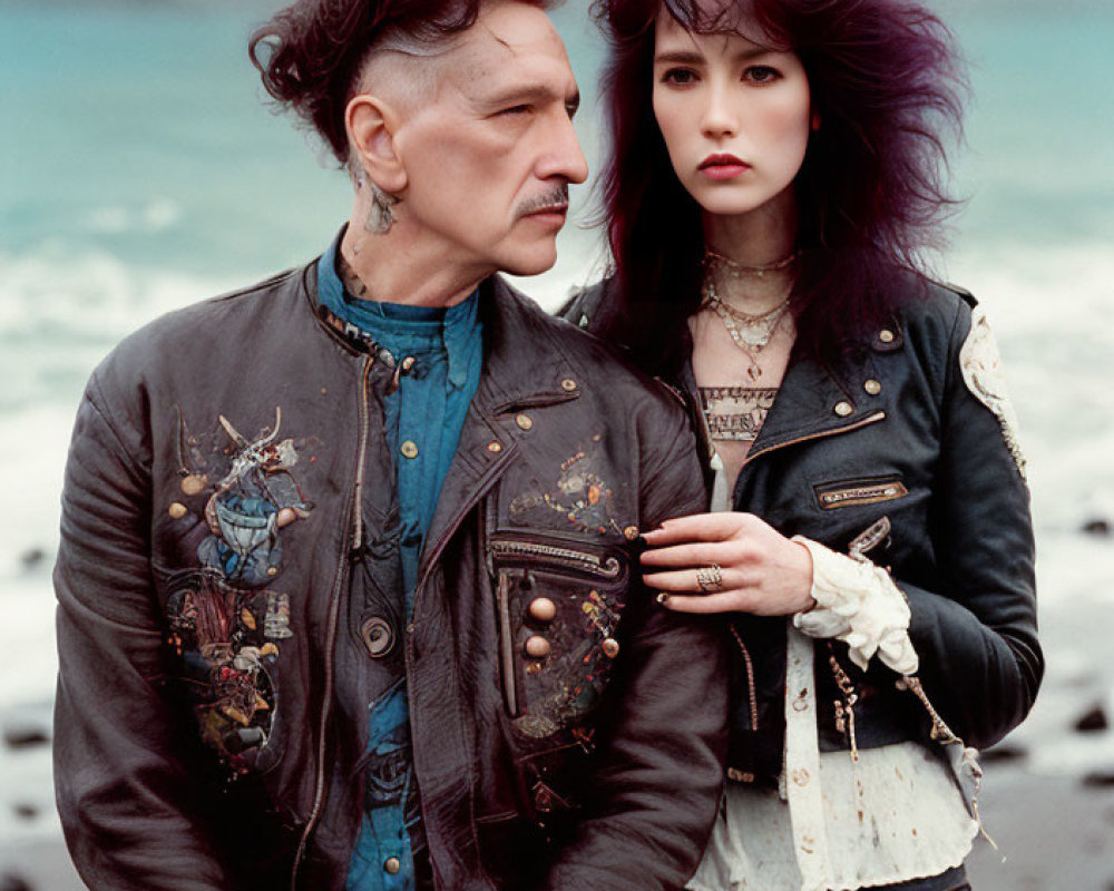 Man and woman in ornate denim jackets pose on pebble beach under cloudy sky