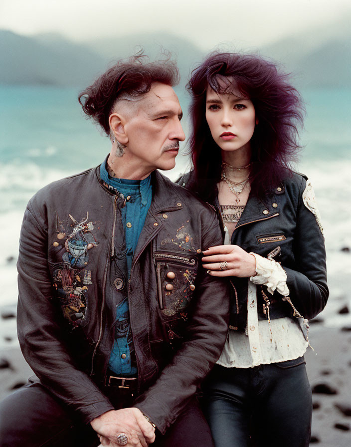Man and woman in ornate denim jackets pose on pebble beach under cloudy sky