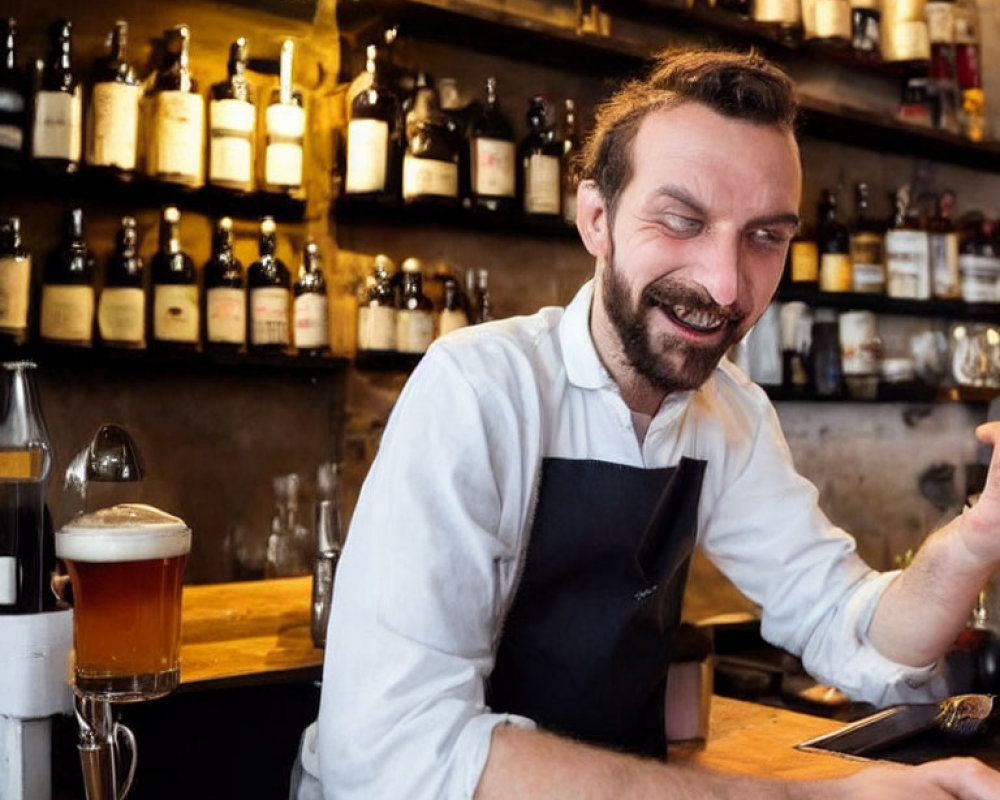 Smiling bartender with beard in apron at bar with bottles and beer