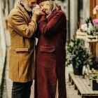 Elderly couple in elegant coats sharing a tender moment by flower-adorned pillar
