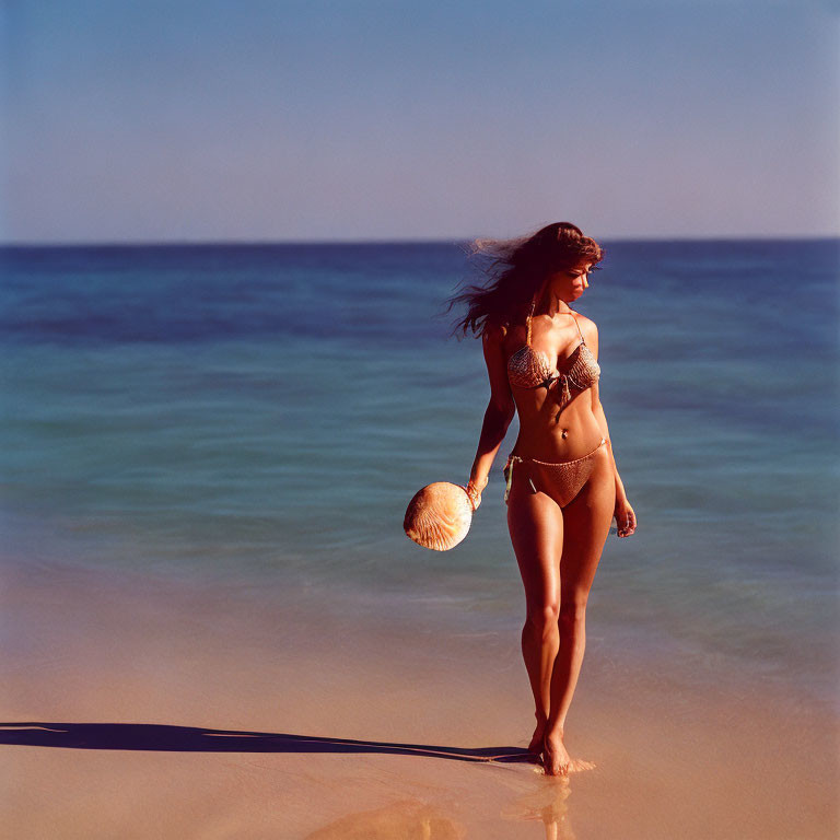 Woman in bikini walking on beach with hat, ocean background & hair blowing