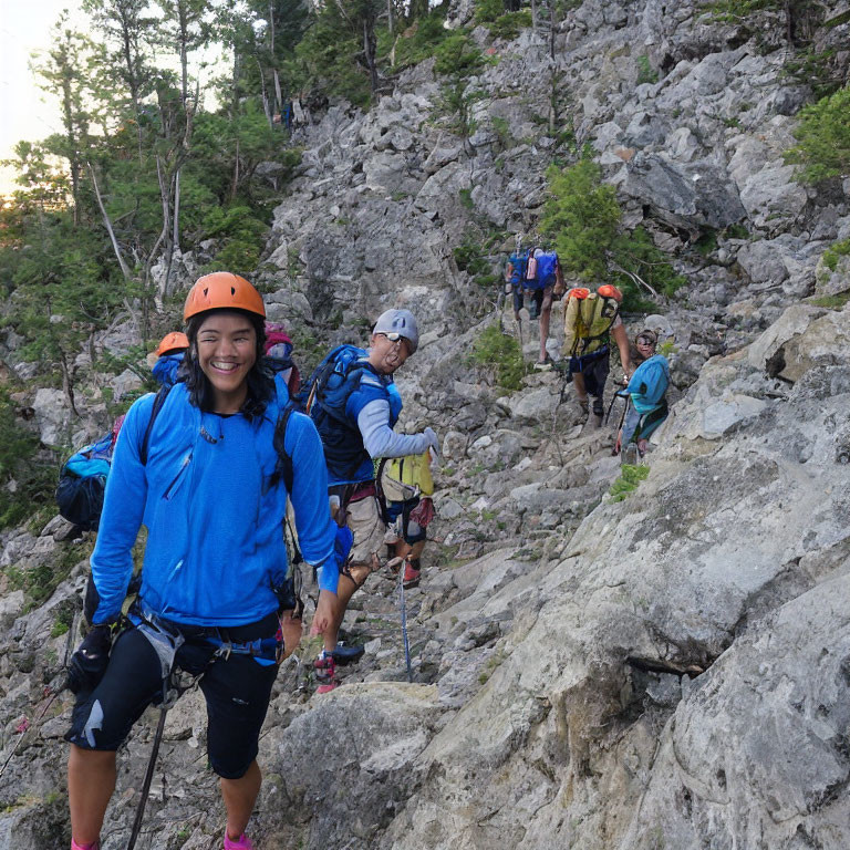 Climbers in safety gear ascending rocky mountain slope with trees in background.