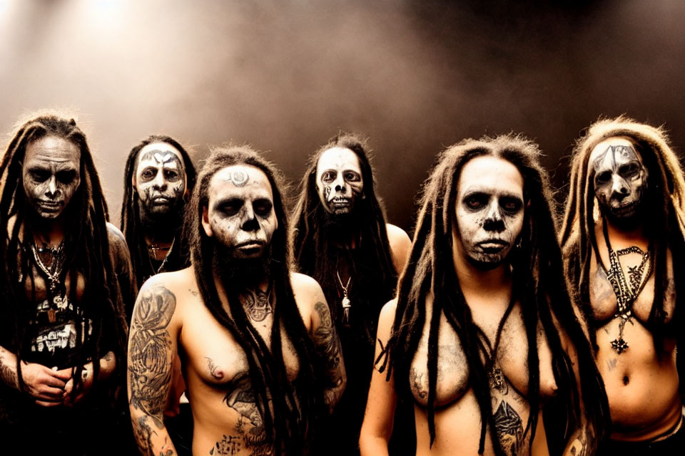 Seven individuals with dark makeup and dreadlocks in intense expressions against a smoky backdrop