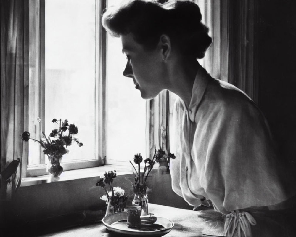 Black-and-white photograph of woman in blouse by window with flowers on table
