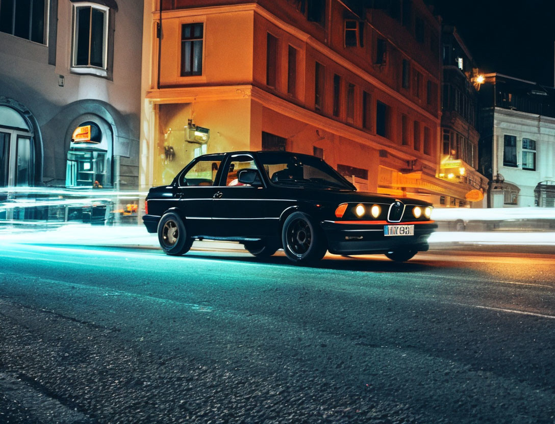 Vintage Black Car Parked on City Street at Night with Passing Vehicle Light Streaks