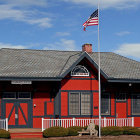 Traditional American schoolhouse with brick walls, porch, flagpole, and figures.