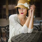Vintage woman in lace dress and tan hat sitting at table, gazing at camera