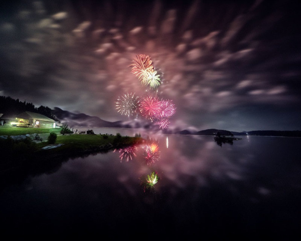 Night sky with fireworks reflecting on tranquil lake, clouds, and hill silhouettes.
