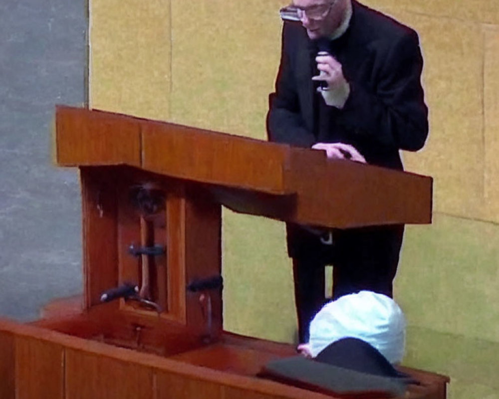 Person in black outfit speaking at wooden podium with seated individual.