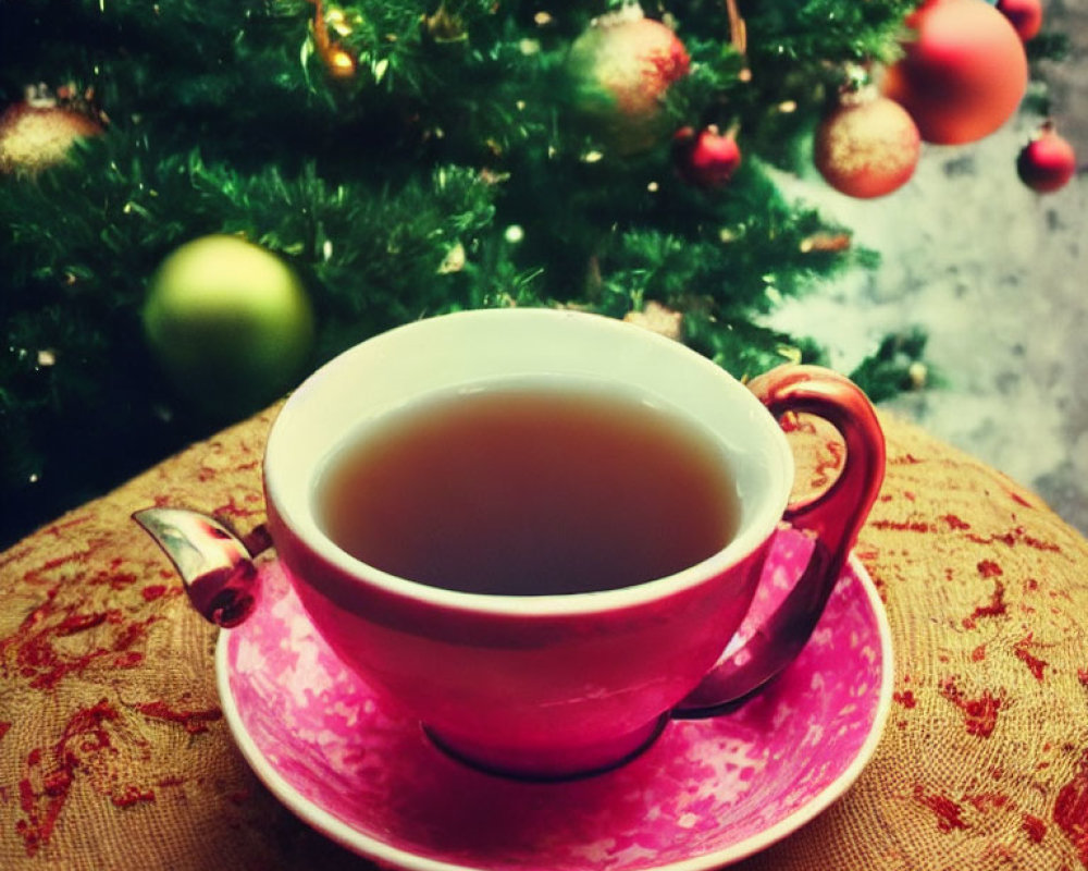 Pink teacup with tea on patterned saucer, blurred Christmas tree with colorful ornaments.