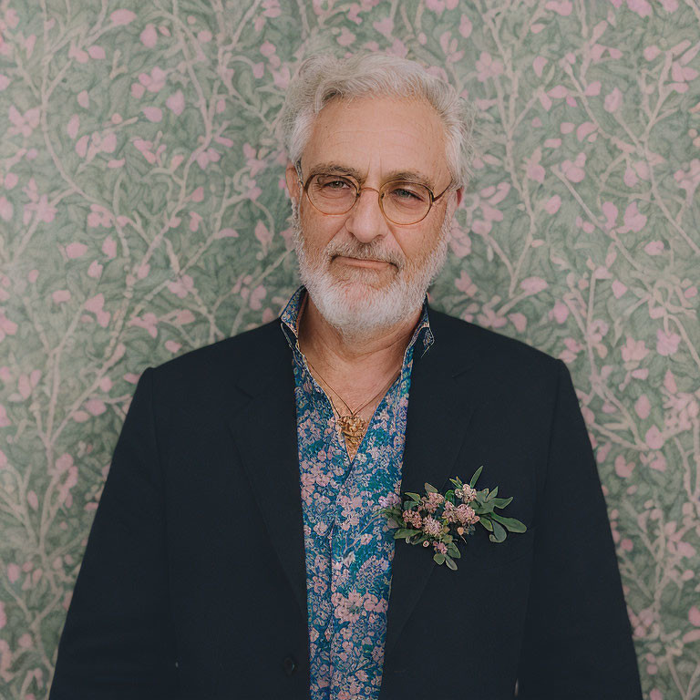 Gray-haired elderly man in glasses, blue shirt, blazer, and floral boutonniere on