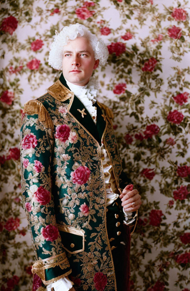 Historical man in green jacket with gold and floral designs against floral wallpaper.