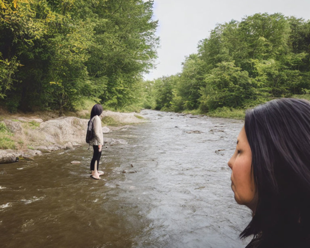 Two people by a river, one in water looking upstream, other onshore surrounded by greenery