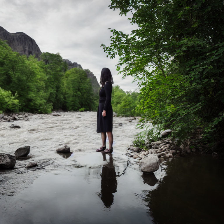 Woman in black dress on rock by river with mountains under cloudy sky