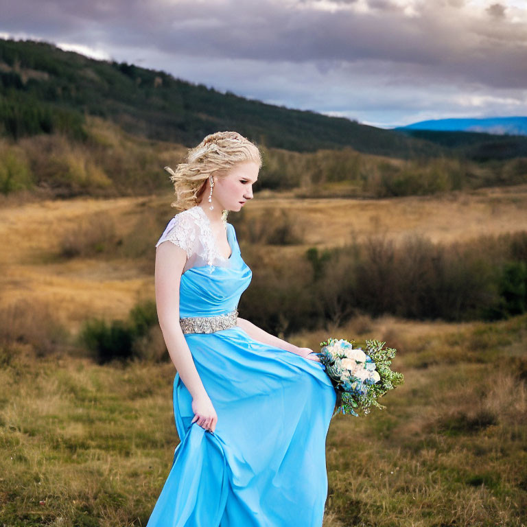 Woman in Blue Dress with Bouquet in Field with Mountains and Cloudy Sky