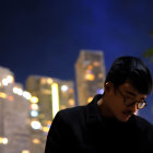 Man in glasses and dark attire stands at night with city lights bokeh.