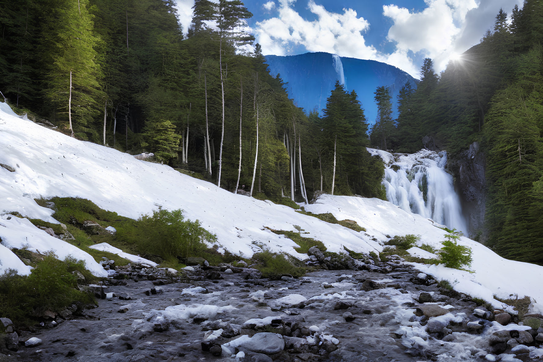 Winter landscape with snowy ground, rocky stream, waterfall, evergreen trees, and sunshine over mountain.